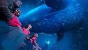 The image shows a family enjoying a realistic underwater projection or exhibit featuring a whale, with expressions of wonder and joy on their faces.