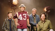 Four men are posing together in front of studio lights, with one wearing a hat and jersey.