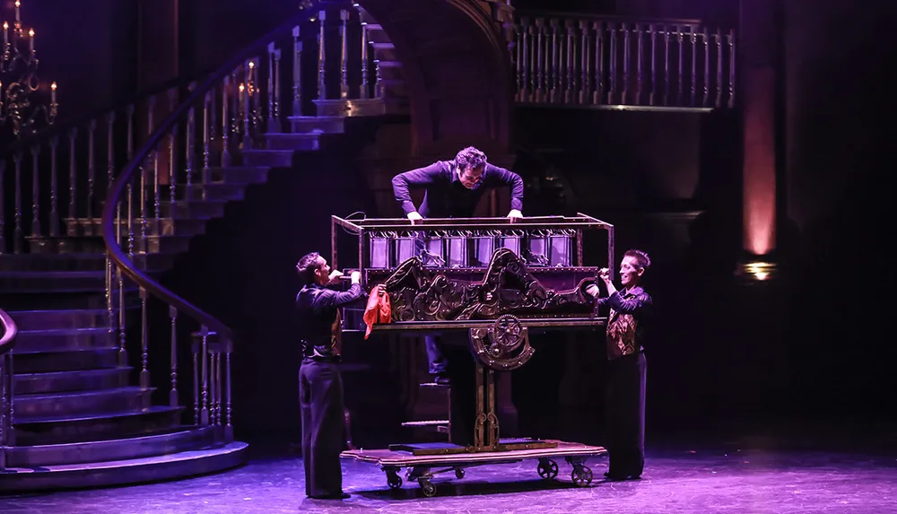 Three performers are seen on stage with one inside a large ornate box in the midst of a magic trick