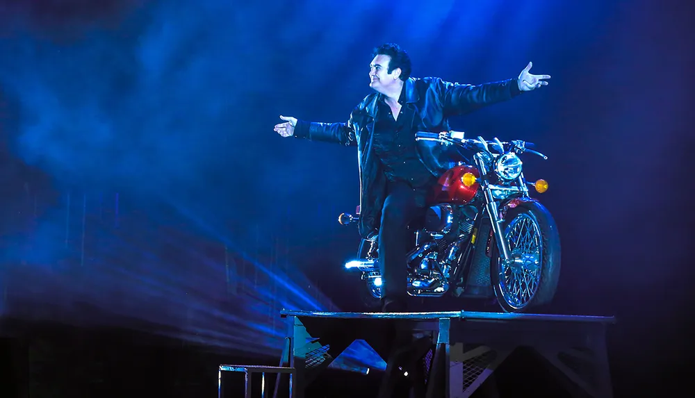 A person in a leather jacket is seated on a motorcycle on a platform striking a dramatic pose under colorful stage lighting