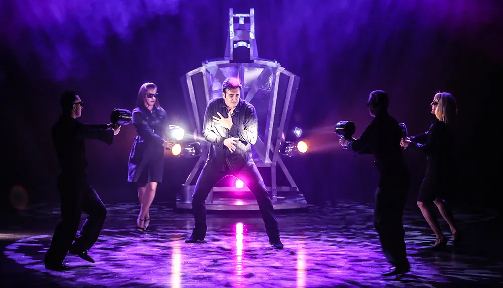 A performer is on stage dramatically clutching their chest surrounded by four individuals pointing flashlights at them in a dimly lit purple-hued atmosphere