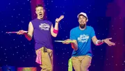 Two performers are on stage with surprised expressions, holding spinning plates on sticks against a starry background.