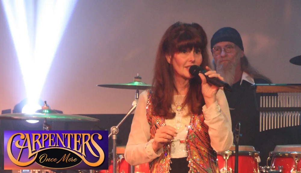 A woman is singing into a microphone on stage with a musician in the background and the overlay text reads CARPENTERS Once More