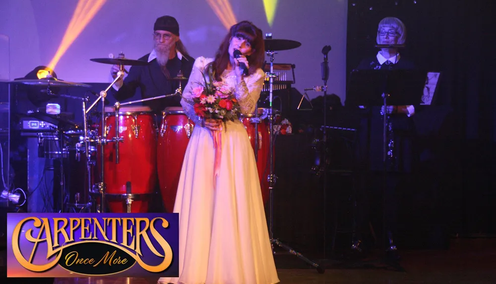A woman in a long white dress sings into a microphone on stage with musicians playing instruments behind her accompanied by a Carpenters Once More logo