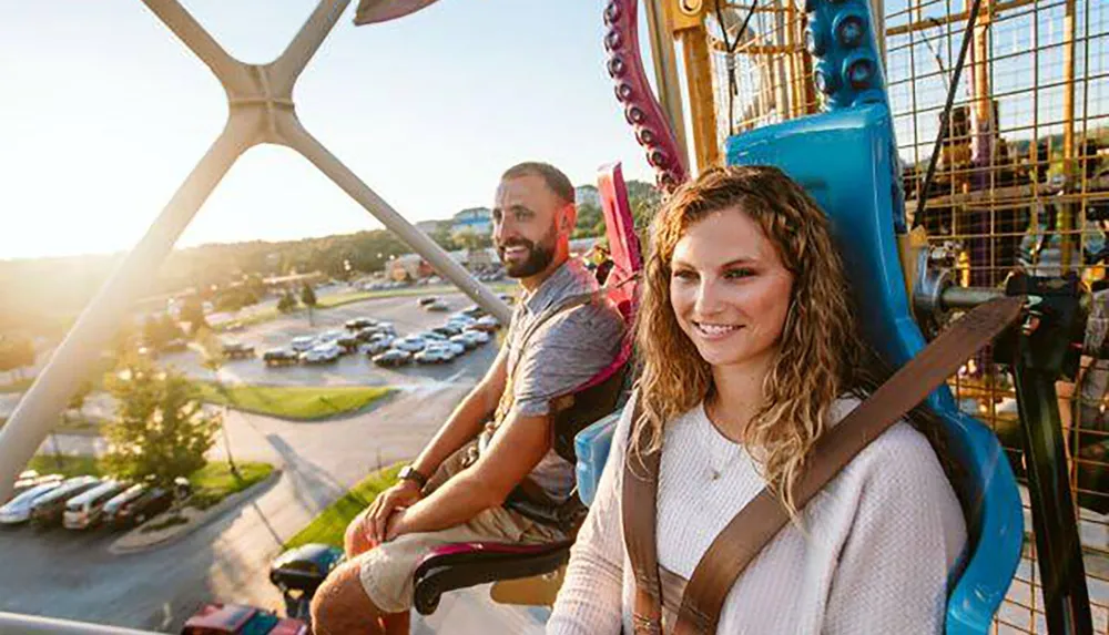 A smiling woman and a man enjoy a ride on a brightly colored Ferris wheel with a view of the surrounding area basked in the glow of the setting sun