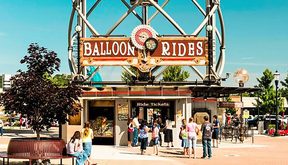 A group of people gathers in front of a balloon ride ticket booth on a sunny day