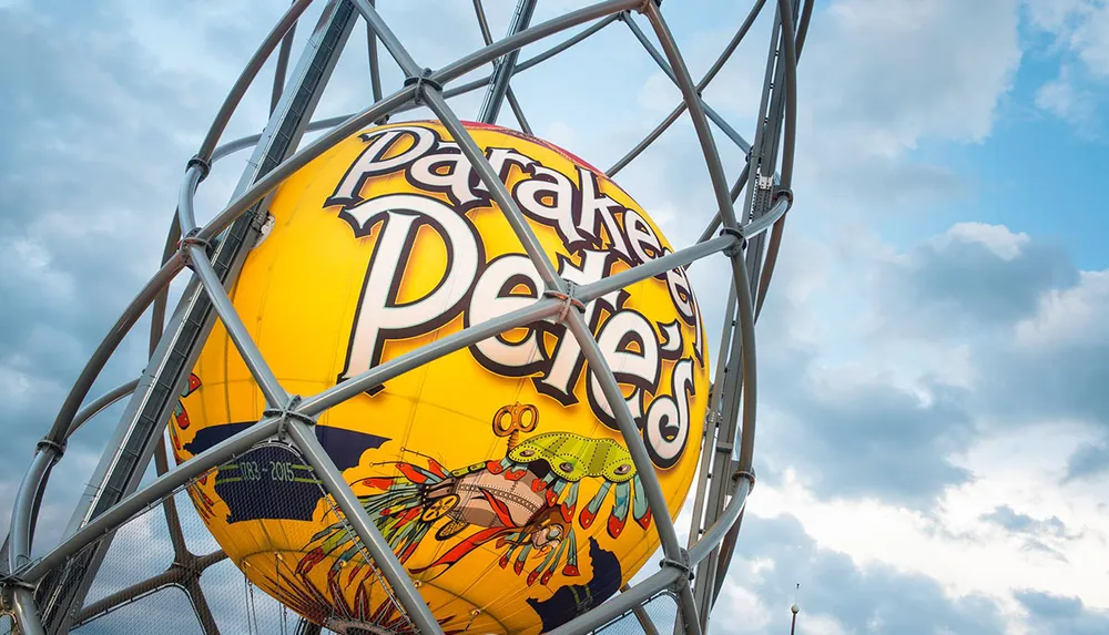 A large yellow sphere with the text Parakeet Petes and colorful graphics is encased in a metal framework against a cloudy sky