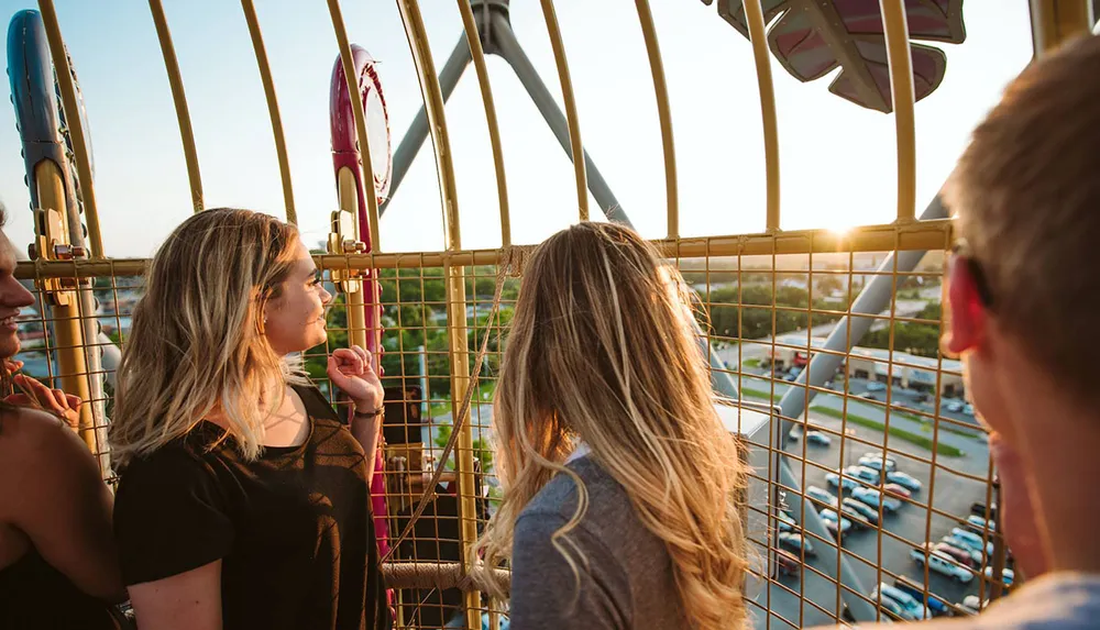 People enjoy the view from high up in a Ferris wheel cage as the sun sets in the background