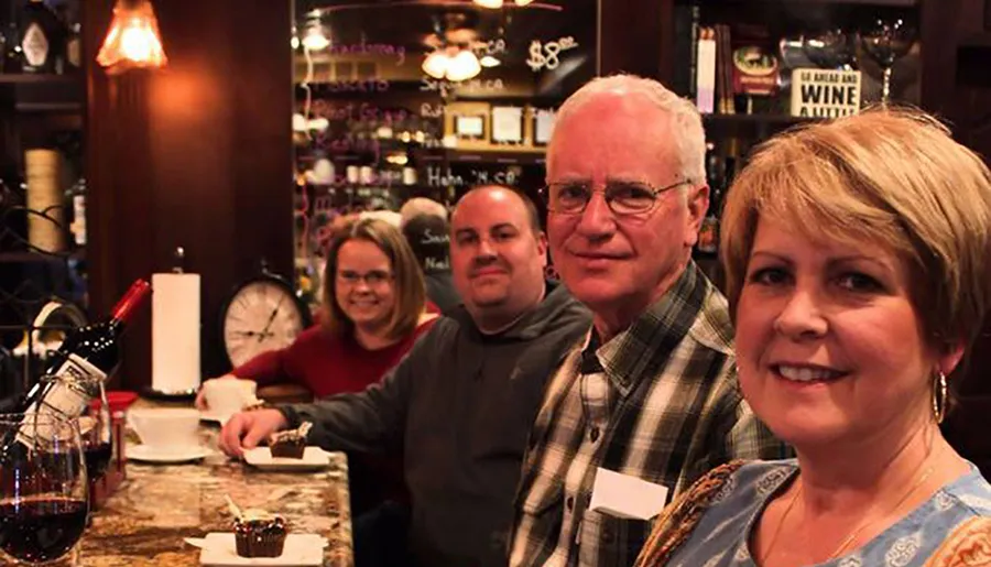 Four people are smiling at the camera in a cozy wine bar setting, with a decanter of red wine and desserts on the counter in the foreground.
