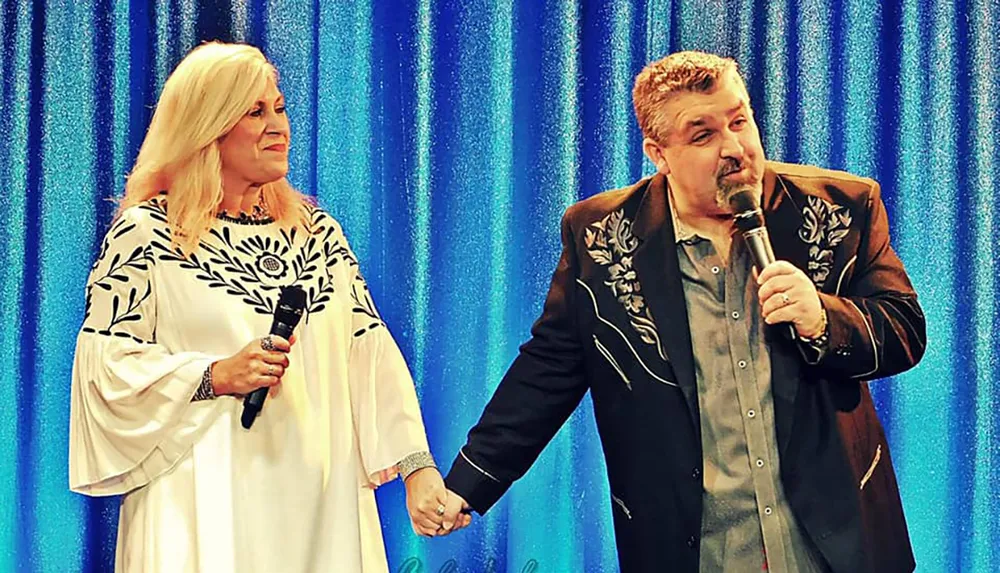 A man and woman both holding microphones and dressed in matching embroidered outfits stand on stage holding hands in front of a blue curtain