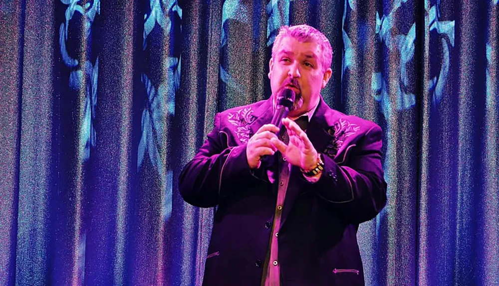 A man is performing on stage with a microphone against a textured dark curtain backdrop with purple and blue lighting