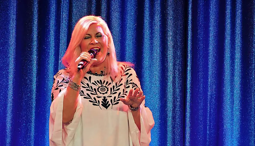 A woman with blonde hair is passionately singing into a microphone in front of a sparkling blue curtain