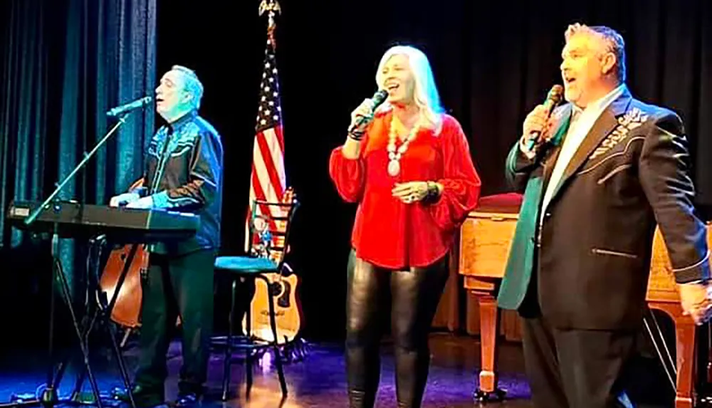 Three people are performing on stage with microphones accompanied by a keyboard and an American flag in the background