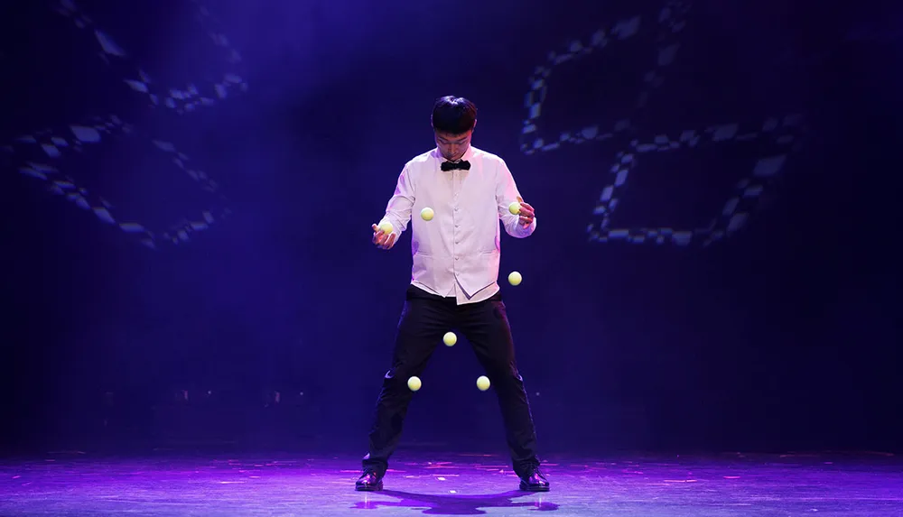 A man in formal attire is juggling multiple balls on a dimly lit stage