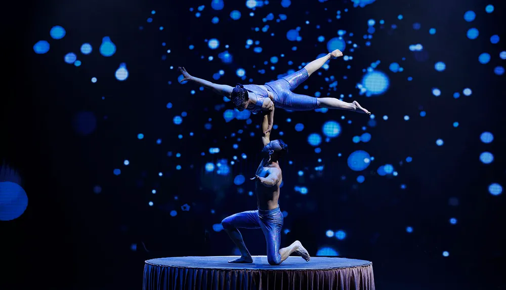 Two acrobats perform a balancing act on a circular stage with a sparkling blue light backdrop