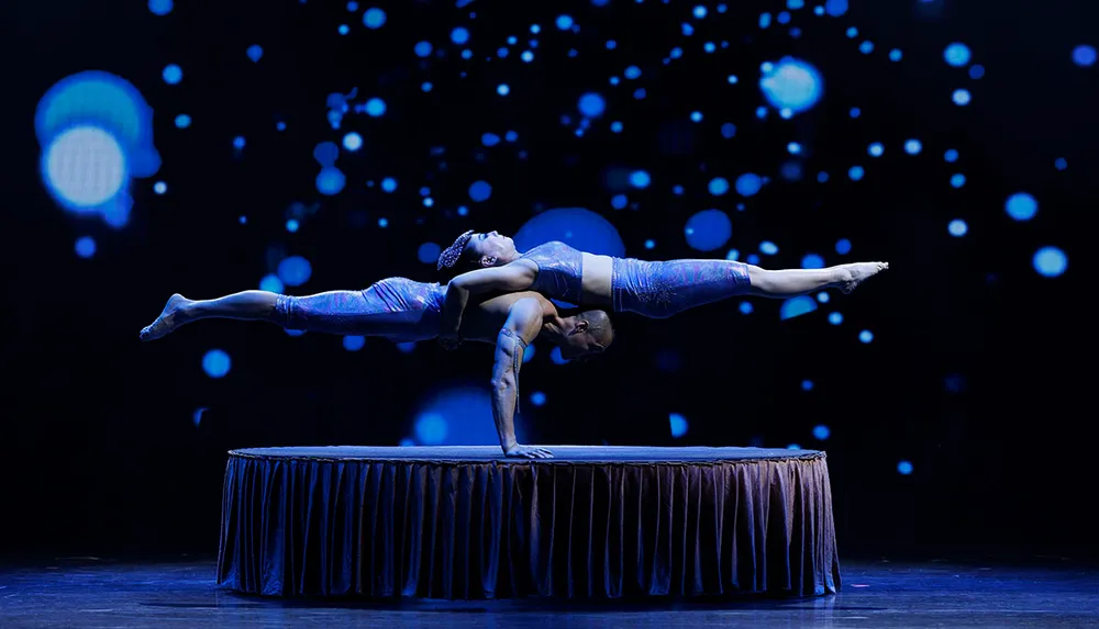A duo of acrobats perform a balancing act with one supporting the other in a horizontal position on a stage with a backdrop of blue bokeh lights