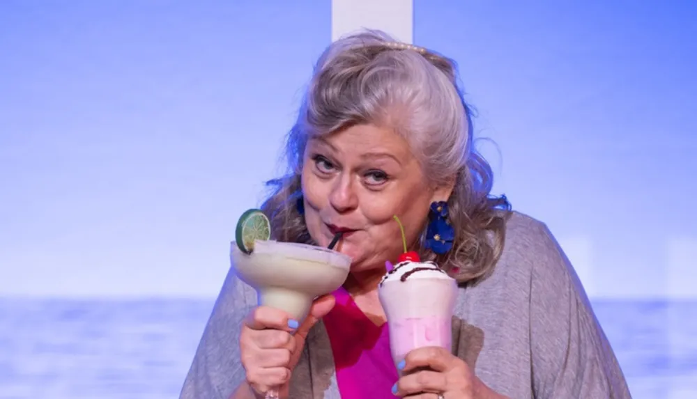 A woman is enjoying two colorful drinks one with a lime garnish and another with whipped cream and a cherry against a blue background