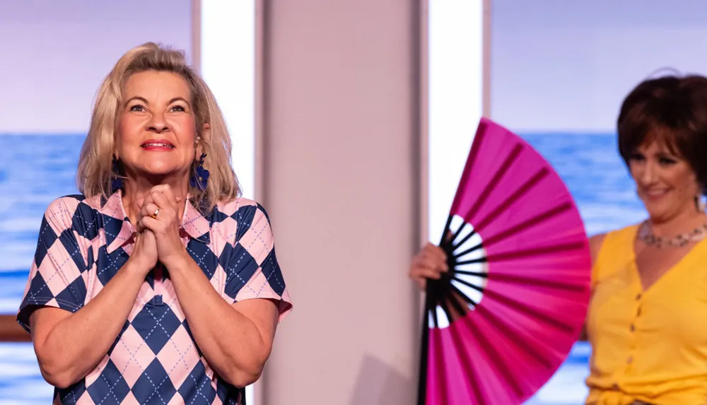 A woman in a pink and blue checkered shirt appears delighted with clasped hands while another woman in a yellow dress holds a large pink fan in the background