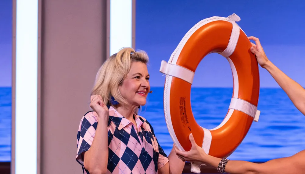 A smiling woman in a patterned shirt is near a person holding an orange lifebuoy with a backdrop of the sea