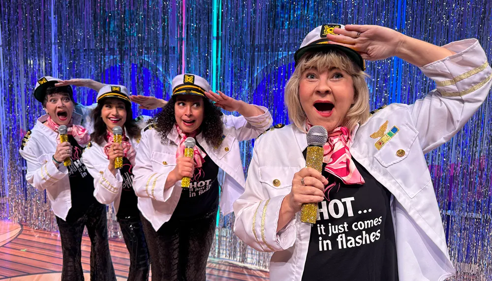 Four women wearing sailor hats and white jackets holding microphones perform enthusiastically in front of a sparkly curtain backdrop
