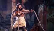 A person is in a theatrical costume, holding a thick rope while striking a determined pose on what appears to be a stage set resembling a rustic environment.