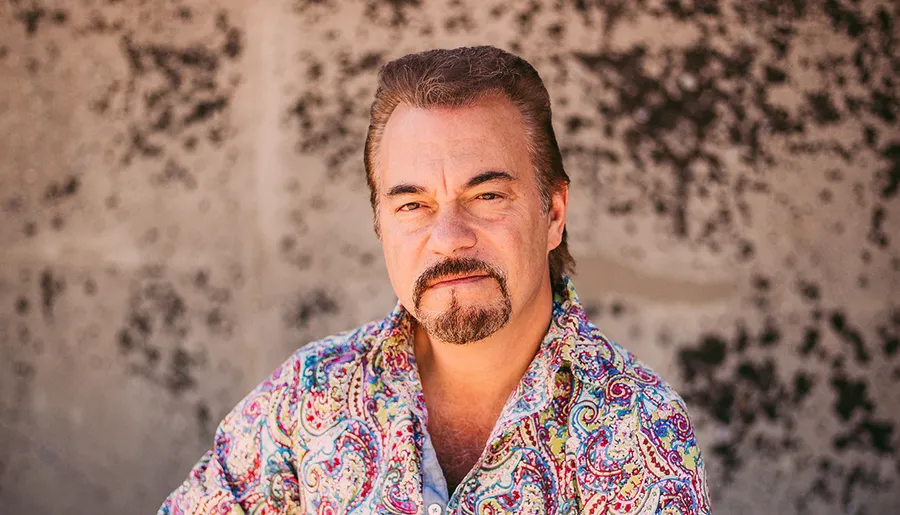 A man with a mustache, wearing a colorful shirt, offers a half-smile against a textured backdrop.