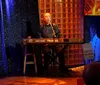 A musician is performing on stage singing into a microphone while playing a Roland RD-700 keyboard set against a backdrop of sparkly blue curtains and decorative panels