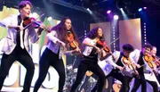 A group of musicians energetically plays violins on stage, wearing matching white jackets, with colorful lights in the background.