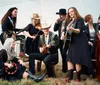 A group of musicians wearing Western-style outfits is performing with various instruments including guitars a violin a mandolin and a double bass in a casual outdoor setting