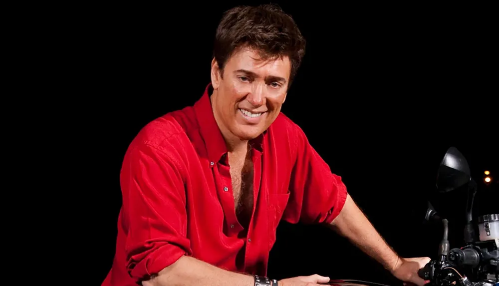 A person with a cheerful smile is dressed in a red shirt and seated on a motorcycle set against a dark background