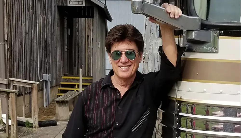 A person with a cheerful expression is standing next to a vehicle wearing sunglasses and holding onto a part of the vehicle with a rustic wooden building in the background