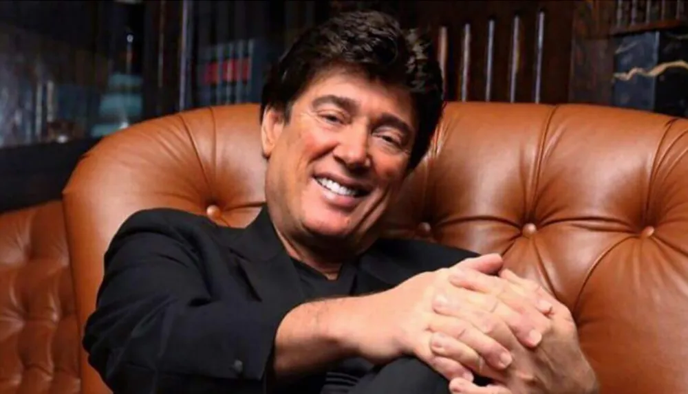 A person is smiling warmly while sitting on a leather sofa with hands clasped together