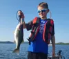 A boy wearing sunglasses and a life vest proudly displays a fish he has caught with a lake and clear sky in the background