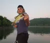 A boy wearing sunglasses and a life vest proudly displays a fish he has caught with a lake and clear sky in the background