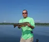 A boy wearing sunglasses and a life vest proudly displays a fish he has caught with a lake and clear sky in the background