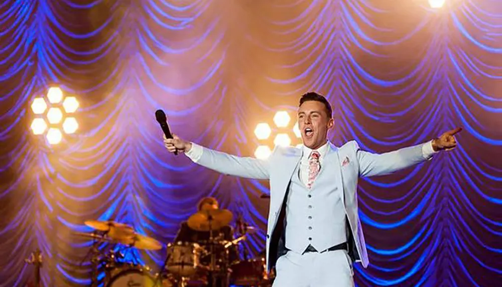 A performer in a white suit with arms outstretched sings into a microphone on a stage with purple lighting and a band in the background