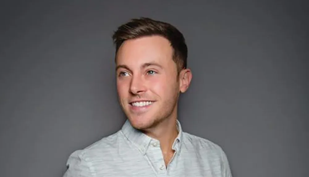 A smiling man in a casual buttoned shirt poses against a neutral gray background