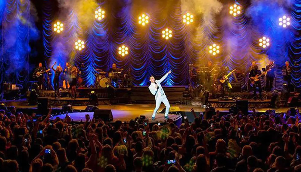 A performer in white is energetically engaging with the crowd during a concert with a live band and a stage lit by warm lights