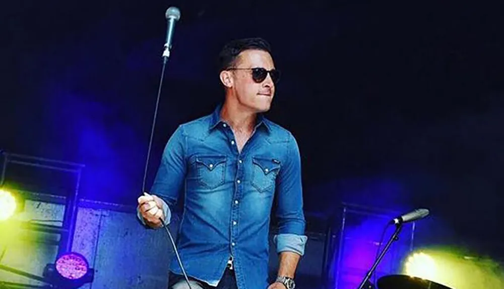 A man in sunglasses and a denim shirt is performing on stage holding onto a microphone stand