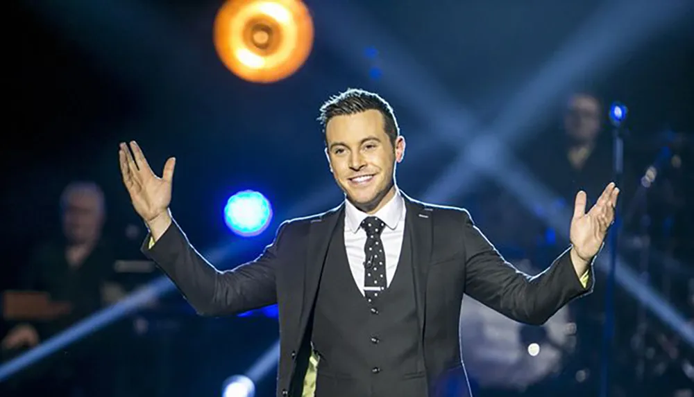 A man in a suit and tie stands on stage with outstretched arms smiling and appearing to engage with an audience under stage lighting