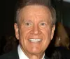 A smiling person is seated with their hands clasped over their knees wearing a blue velvet jacket and paisley tie with books and a vintage television set in the background