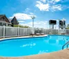 The image shows a clear blue swimming pool surrounded by a white fence with a building and a sign visible in the background under a bright blue sky with scattered clouds