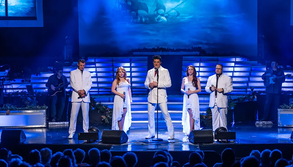 A group of performers dressed in white stands on a stage with blue lighting singing into microphones before an audience