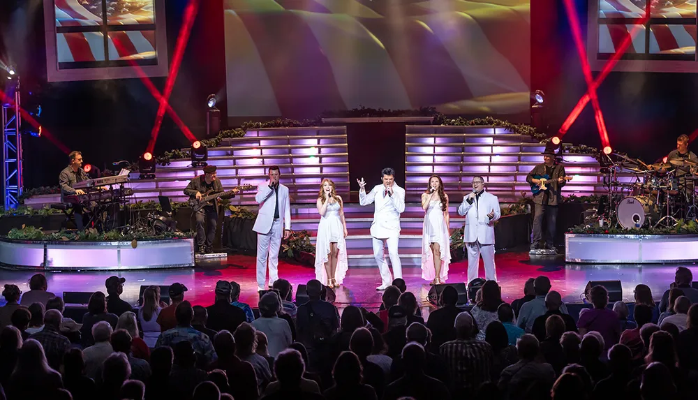 A group of performers dressed in white are singing on a brightly lit stage in front of an audience