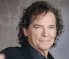 The image shows a confident middle-aged man with curly hair wearing a black blazer posing for the camera against a textured grey background