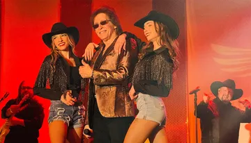 A performer wearing a shiny jacket poses on stage with two women in hats and fringe outfits, set against a red background.