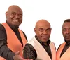 Three men in matching black and orange vests are posing together for a photo with one slightly in front of the other two all looking directly at the camera