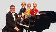 A smiling man in glasses and a tuxedo is seated at a grand piano with a Boston Terrier on the keyboard, flanked by three lifelike puppets and a rabbit perched in a top hat.