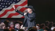 A performer in a cowboy hat is singing into a microphone with an American flag backdrop.