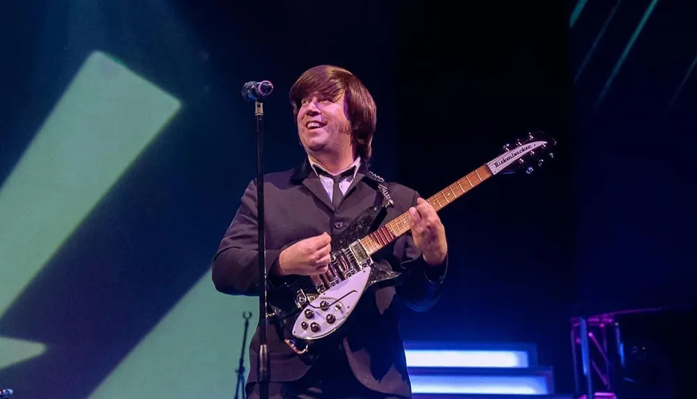 A musician in a suit is playing a Rickenbacker guitar on stage smiling while performing